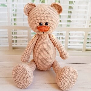 May include: A crocheted beige teddy bear with orange nose and black eyes, sitting on a white wooden surface.