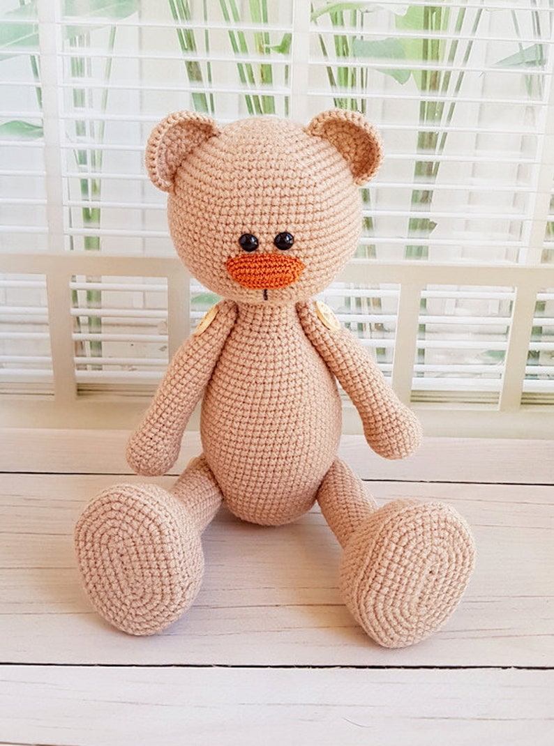 May include: A crocheted beige teddy bear with orange nose and black eyes, sitting on a white wooden surface.