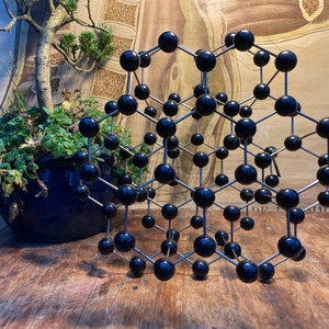 May include: A geometric sculpture featuring black spheres joined by silver rods. It rests on a wooden surface, with a potted plant and a vintage anatomical chart in the background. The sculpture is a decorative item.