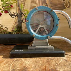 May include: An antique scientific instrument with a circular, light blue frame and a clear glass window. The instrument has a silver-colored base and curved handles. A small bonsai tree with red berries is in the background.