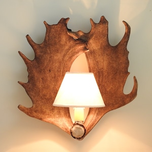 May include: A wall-mounted lamp made from a pair of antlers with a white lampshade. The antlers are brown and have a natural finish.