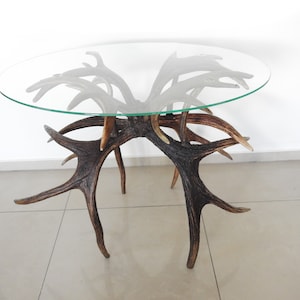 May include: A round glass table top sits atop a base made of multiple antlers. The antlers are arranged in a radial pattern, with the points of the antlers facing outwards. The table is made of natural materials and has a rustic, organic look.