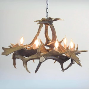 Peut inclure: Un lustre composé de plusieurs bois de cerf bruns et texturés disposés en cercle. Le luminaire comporte cinq ampoules verticales en forme de bougie avec des filaments à lueur chaude, suspendu par une chaîne en métal noir au sommet central.