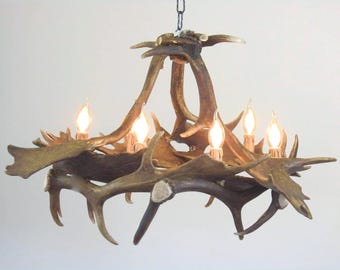 Può includere: Un lampadario costruito con molteplici palchi di cervo marroni e testurizzati disposti in una cornice circolare. L'apparecchio presenta cinque lampadine verticali a forma di candela con filamenti a luce calda, sospeso da una catena in metallo nero.