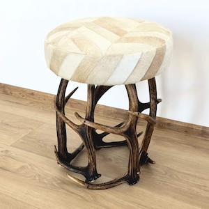 May include: A round stool featuring a seat upholstered in a chevron-patterned cowhide with shades of cream, beige, and white. The base is constructed from several dark brown, textured antlers arranged in a circular formation to support the seat.