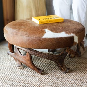 May include: Round ottoman with a cowhide top in shades of brown and white. The base is made of intricately carved wood, resembling antlers. A yellow book rests on the ottoman. The ottoman is approximately 18 inches in diameter.