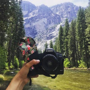 May include: A black Canon EOS Mark II camera with a floral camera strap, held in front of a river and mountains.