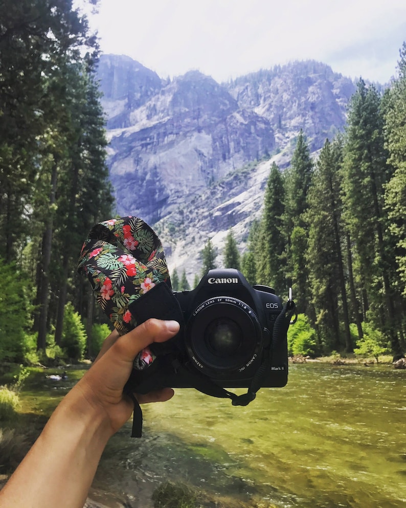 May include: A black Canon EOS Mark II camera with a floral camera strap, held in front of a river and mountains.