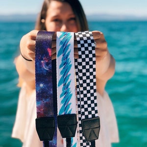Op de afbeelding: Vier camera-riemen met verschillende patronen: galaxy print, blauw-wit geometrisch patroon, zwart-wit geruit patroon en een effen zwarte riem.