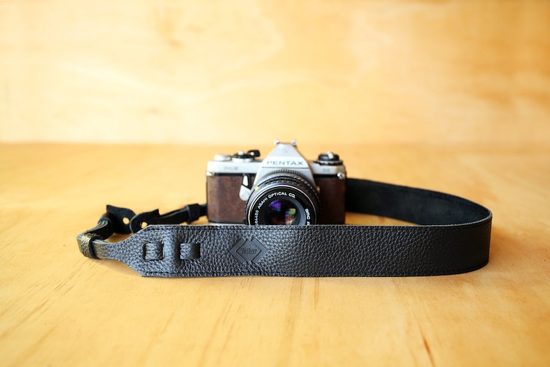 May include: A black leather camera strap with a silver metal buckle and the word "Artisan" embossed on the strap. The strap is attached to a silver camera with a black lens.