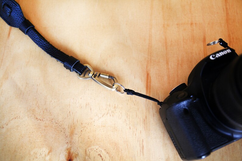 May include: A black leather camera strap with a silver carabiner clip attached to a black Canon camera.