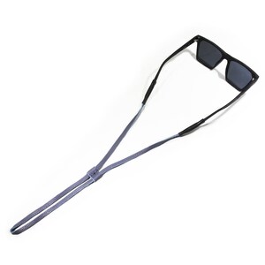 K&ouml;nnte beinhalten: Eine schwarze Sonnenbrille mit einem grauen Lederband. Das Band ist verstellbar und kann um den Hals getragen werden.