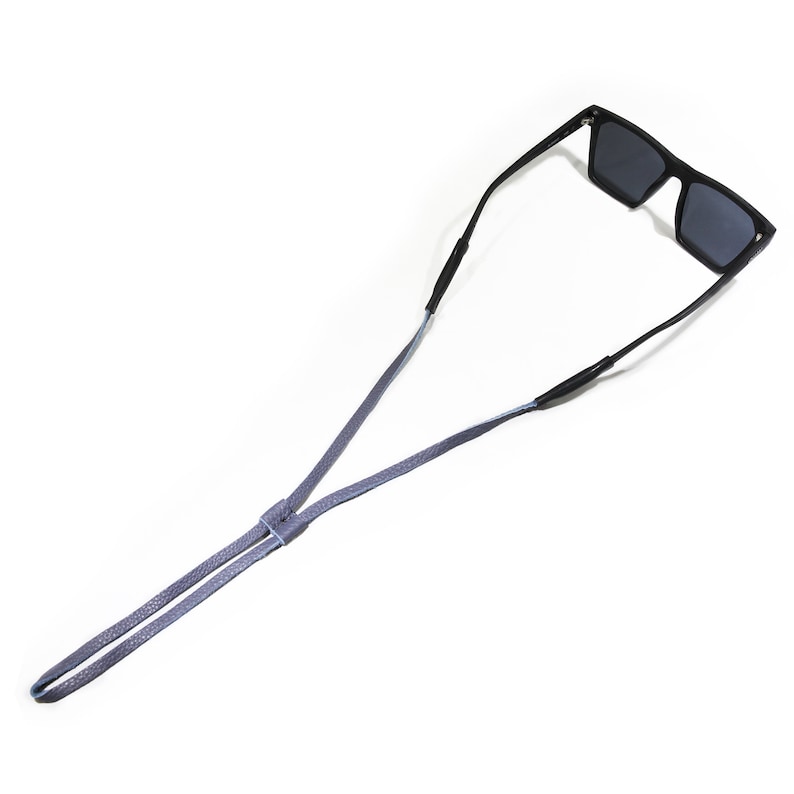 K&ouml;nnte beinhalten: Eine schwarze Sonnenbrille mit einem grauen Lederband. Das Band ist verstellbar und kann um den Hals getragen werden.