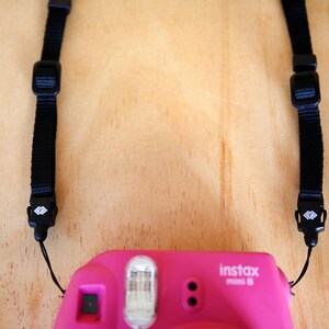May include: A pink instant camera with a black strap. The camera has a flash and a lens. The strap has a diamond-shaped logo on the buckles.