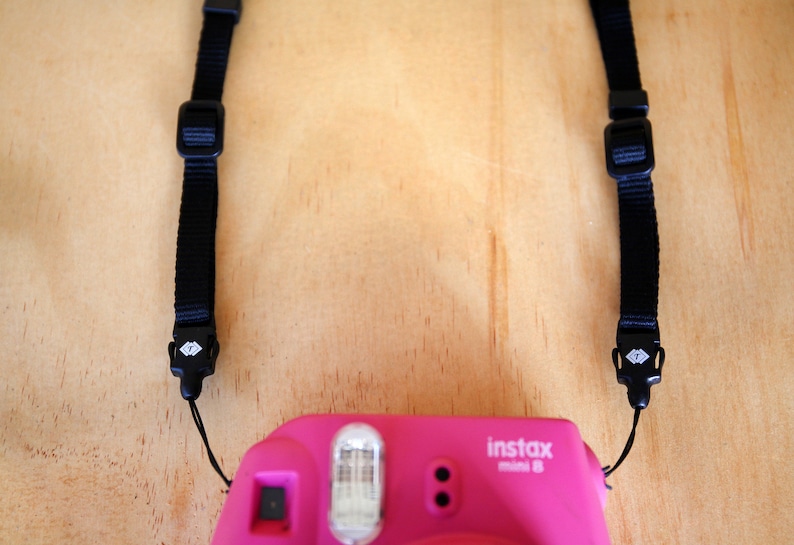 May include: A pink instant camera with a black strap. The camera has a flash and a lens. The strap has a diamond-shaped logo on the buckles.