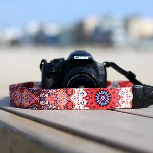 Op de afbeelding: Een zwarte Canon camera met een gepatenteerde cameraband. De band is rood, oranje, blauw en wit met een mandala-ontwerp.