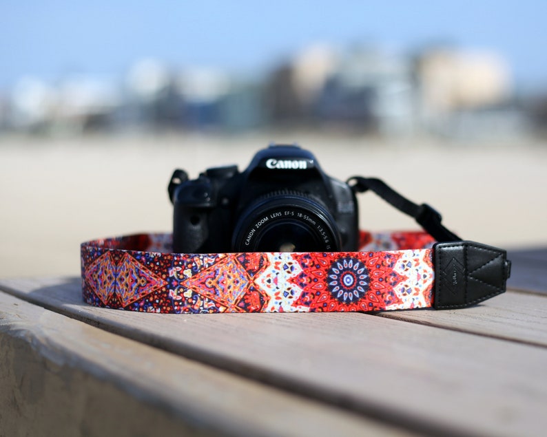 Op de afbeelding: Een zwarte Canon camera met een gepatenteerde cameraband. De band is rood, oranje, blauw en wit met een mandala-ontwerp.