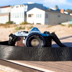 May include: A black leather camera strap with the word "Camera" embossed on it. The strap is attached to a silver camera lens with the text "Asahi Optical Co. Pentax" printed on it.
