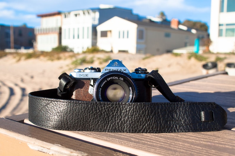 May include: A black leather camera strap with the word "Camera" embossed on it. The strap is attached to a silver camera lens with the text "Asahi Optical Co. Pentax" printed on it.