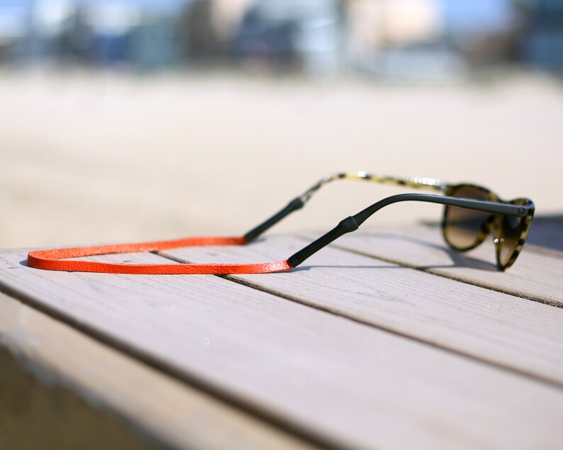Peut inclure: Une paire de lunettes de soleil avec une monture noire et des verres bruns, pos&eacute;e sur une surface en bois. Un cordon en cuir orange vif est attach&eacute; aux lunettes.