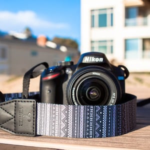 Op de afbeelding: Een zwarte Nikon DSLR-camera met een lens erop, die op een houten oppervlak ligt. De camera is bevestigd aan een zwart-witte gepatenteerde cameraband met het woord "Tether" erop gedrukt.
