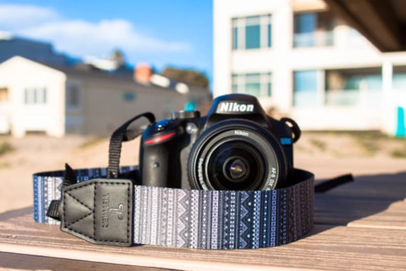 Op de afbeelding: Een zwarte Nikon DSLR-camera met een lens erop, die op een houten oppervlak ligt. De camera is bevestigd aan een zwart-witte gepatenteerde cameraband met het woord "Tether" erop gedrukt.