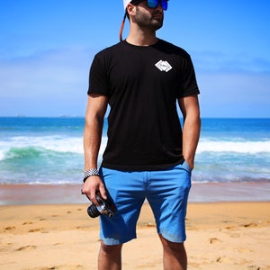 May include: A man wearing a black t-shirt with a white logo that says "Cove" on it, blue shorts, and sunglasses. He is standing on a sandy beach with the ocean in the background.