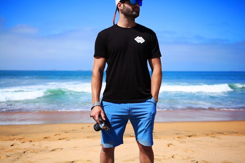 May include: A man wearing a black t-shirt with a white logo that says "Cove" on it, blue shorts, and sunglasses. He is standing on a sandy beach with the ocean in the background.