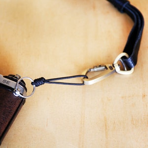 May include: A black camera strap with a silver carabiner clip attached to a black and silver camera. The camera has a silver lens and a brown leather body. The strap is made of a woven material and has a knot near the camera.