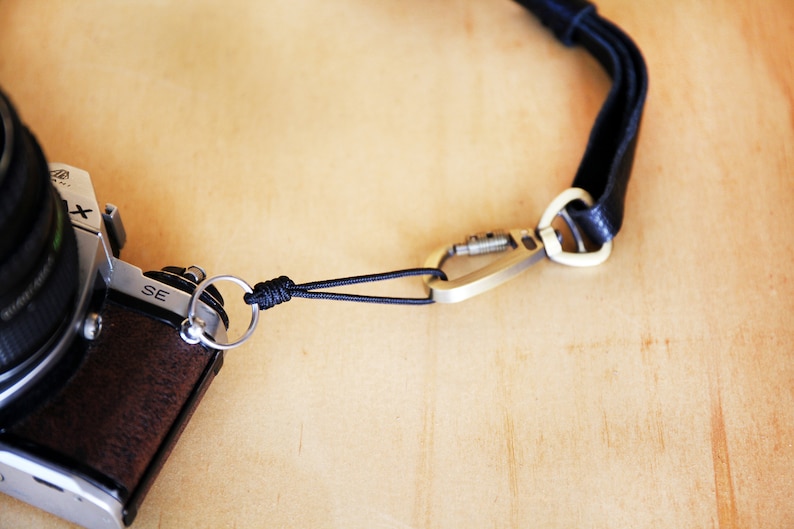 May include: A black camera strap with a silver carabiner clip attached to a black and silver camera. The camera has a silver lens and a brown leather body. The strap is made of a woven material and has a knot near the camera.