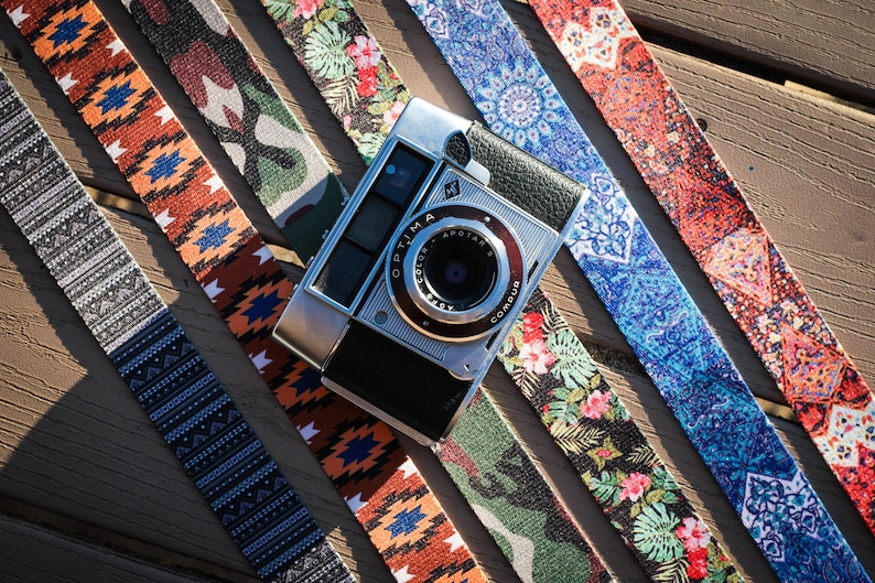 Op de afbeelding: Een zilveren camera met een zwarte leren band en een lens met de woorden "OPTIMA" en "APOTAR" erop gedrukt. De camera ligt op vijf kleurrijke, gepatroneerde camerabandjes.