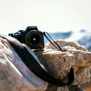 May include: A black Canon camera with a lens and a black strap is resting on a white and brown rock.