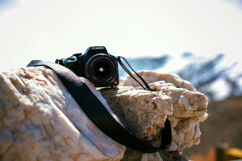 May include: A black Canon camera with a lens and a black strap is resting on a white and brown rock.