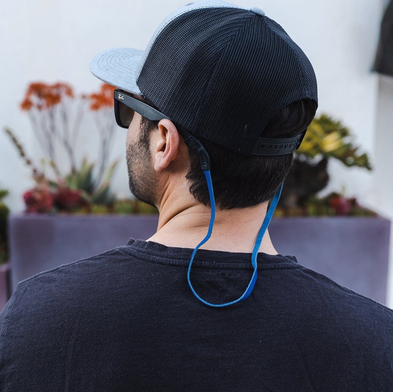 Peut inclure: Une personne portant une casquette de baseball noire et grise avec une sangle bleue. La sangle est pass&eacute;e autour de la nuque. La personne porte des lunettes de soleil.