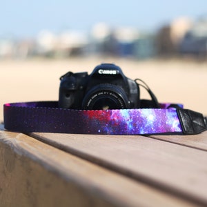 Op de afbeelding: Een zwarte DSLR-camera met een camera-band met een galaxy-thema. De band is gemaakt van een donkere stof met een paarse, blauwe en roze galaxy-print.