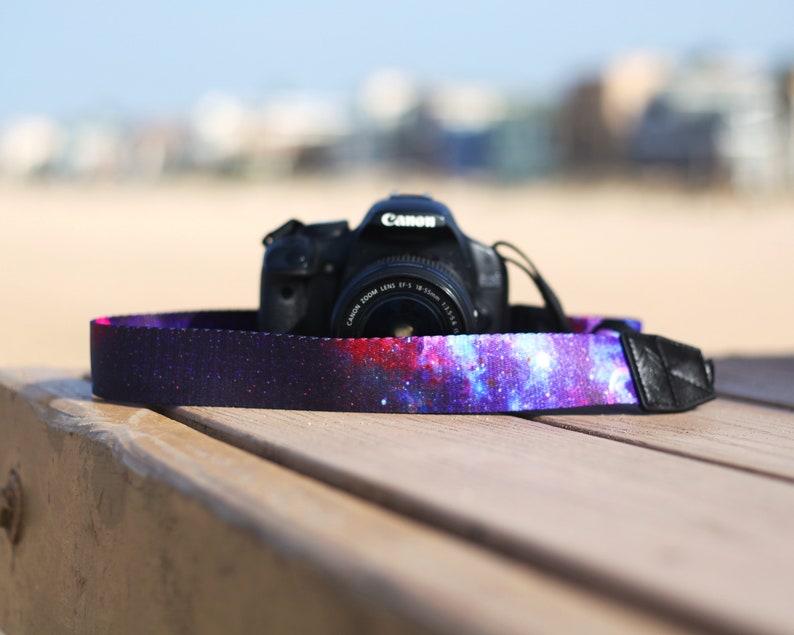 Op de afbeelding: Een zwarte DSLR-camera met een camera-band met een galaxy-thema. De band is gemaakt van een donkere stof met een paarse, blauwe en roze galaxy-print.