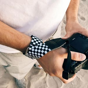 May include: A black and white checkered wrist strap for a camera. The strap is attached to a black camera with a black lens.