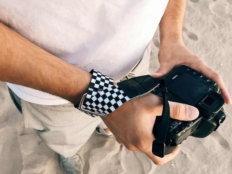 May include: A black and white checkered wrist strap for a camera. The strap is attached to a black camera with a black lens.