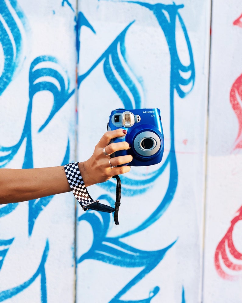 May include: A blue Polaroid 300 instant camera with a black and white checkered camera strap. The camera is held in front of a white wall with blue and red graffiti.