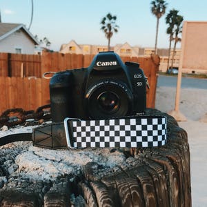 May include: A black Canon EOS 5D Mark II camera with a black and white checkered camera strap. The camera is sitting on a weathered wooden surface.