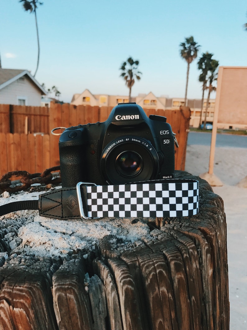 May include: A black Canon EOS 5D Mark II camera with a black and white checkered camera strap. The camera is sitting on a weathered wooden surface.