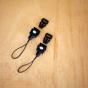 May include: Two black plastic clasps with a small white logo on each. The clasps are attached to black cords with loops at the end.