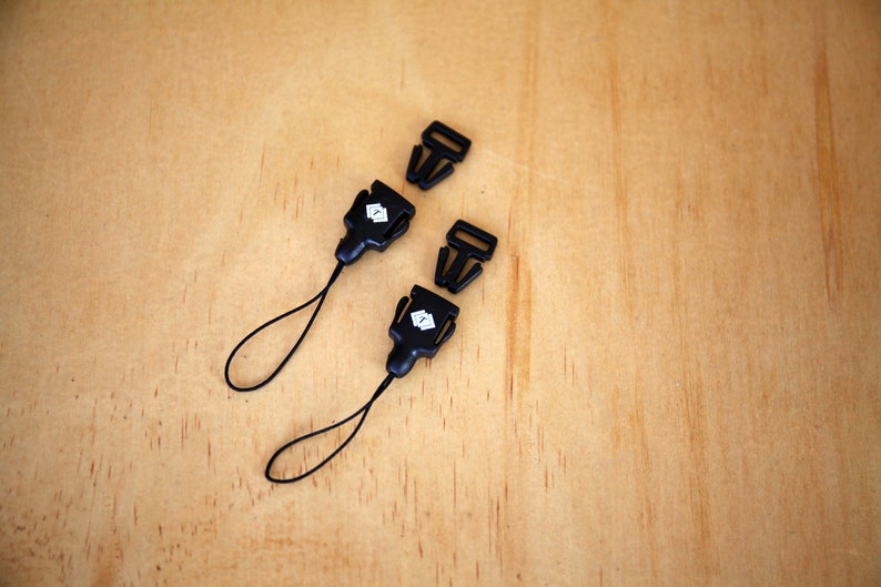 May include: Two black plastic clasps with a small white logo on each. The clasps are attached to black cords with loops at the end.
