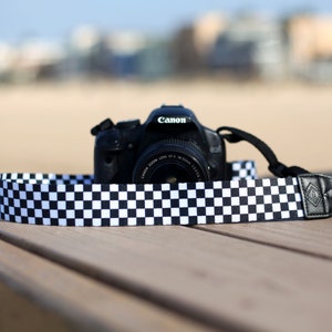 Op de afbeelding: Een zwarte Canon-camera met een geruite cameraband. De camera ligt op een houten oppervlak.