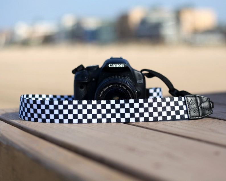Op de afbeelding: Een zwarte Canon-camera met een geruite cameraband. De camera ligt op een houten oppervlak.