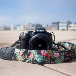 Op de afbeelding: Een zwarte DSLR-camera met een camera-riem met bloemenpatroon. De camera lens is een Nikon AF-S DX NIKKOR 18-55mm f/3.5-5.6G VR.