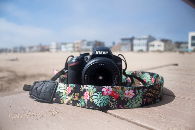 Op de afbeelding: Een zwarte DSLR-camera met een camera-riem met bloemenpatroon. De camera lens is een Nikon AF-S DX NIKKOR 18-55mm f/3.5-5.6G VR.