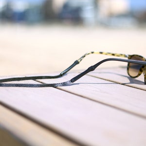 Peut inclure: Un cordon en cuir noir pour lunettes de soleil, avec les lunettes pos&eacute;es sur une surface en bois.