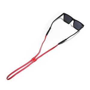 K&ouml;nnte beinhalten: Ein rotes Lederband f&uuml;r Sonnenbrillen. Das Band besteht aus zwei Lederst&uuml;cken, die in der Mitte miteinander verbunden sind. Das Band ist lang genug, um um den Hals getragen zu werden.