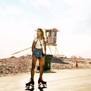 May include: A woman wearing a white crop top and denim shorts is roller skating on a paved path. She is wearing a camera strap around her neck and has a lifeguard stand in the background.